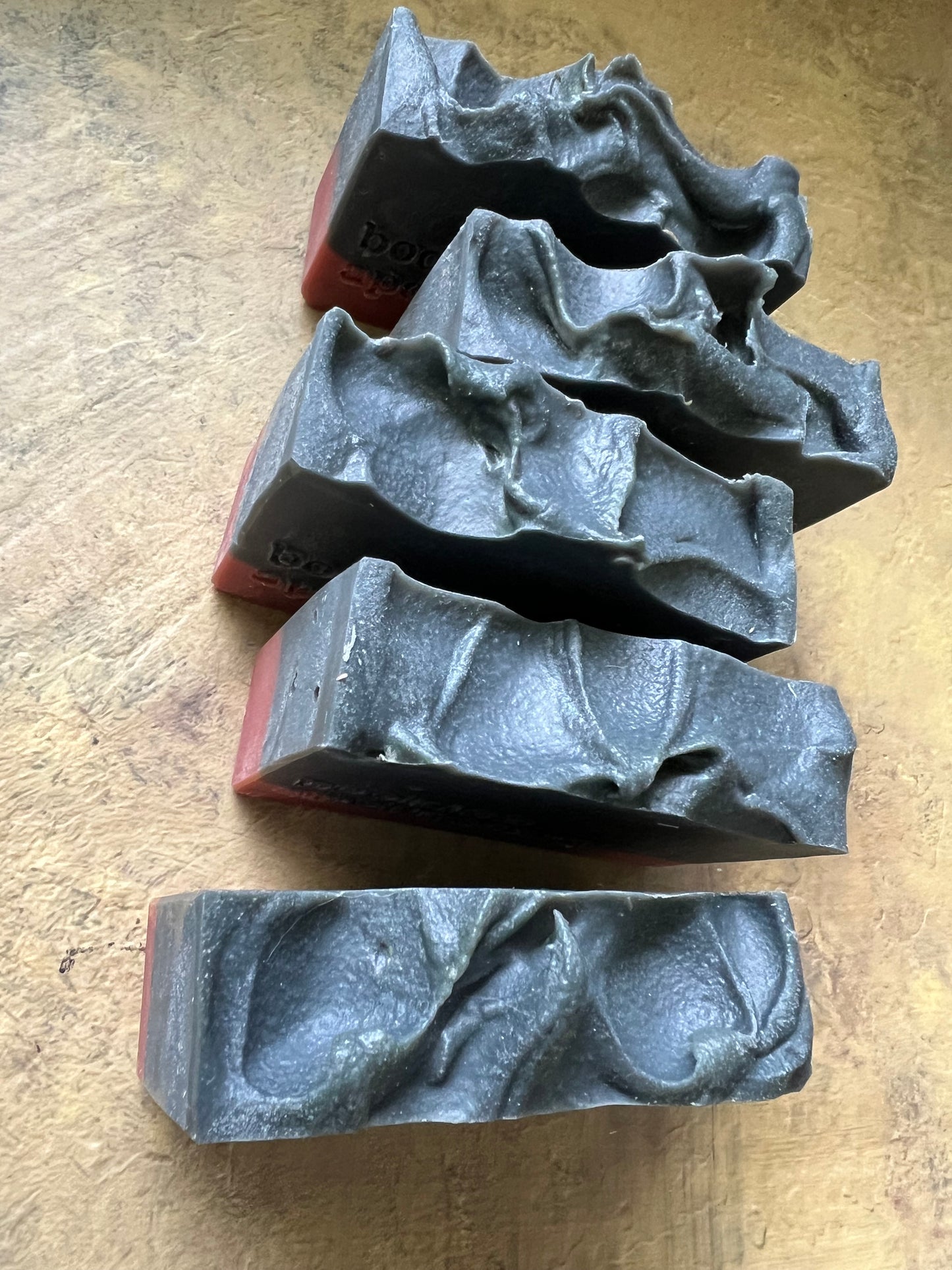 Five dark gray, textured rectangular bars with wavy, organic patterns on top, arranged on a textured tan surface.