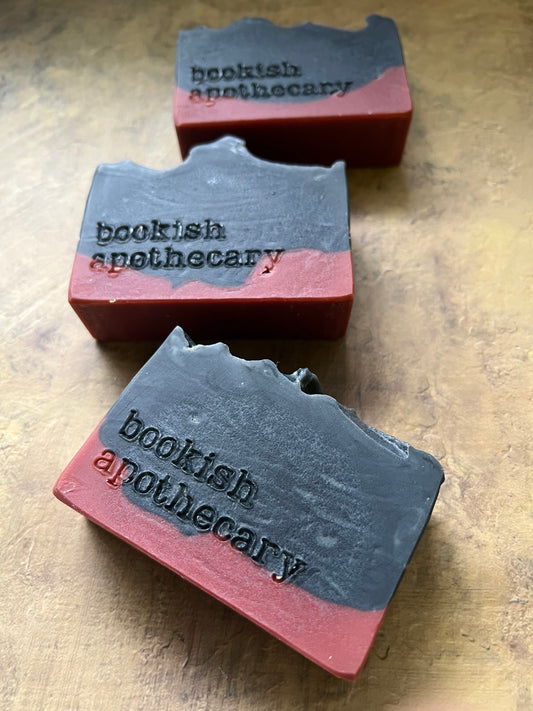 Three handmade soap bars with a dark gray top and a dusty rose pink base, each hand-stamped with the words 'bookish apothecary'. They are arranged on a textured, warm-toned wooden surface.