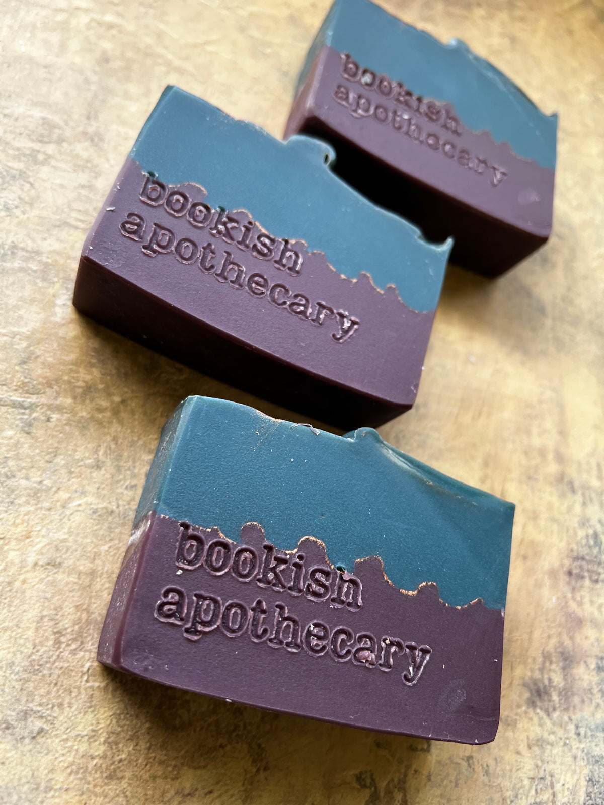 Three rectangular bars of handmade soap with a two-tone design, featuring a dark teal top half and a deep plum-purple bottom half embossed with the brand name 'bookish apothecary', arranged on a textured brown surface.