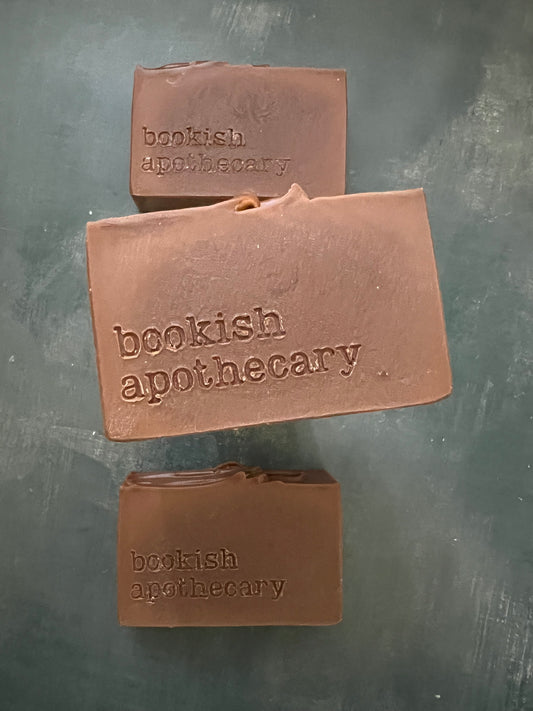 Three bars of gold soap on a dark gray textured surface, each stamped with the words 'bookish apothecary'.