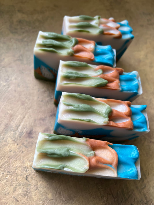 Five handmade soap bars with a layered, swirled pattern of blue, peach, green, and white, arranged on a textured brown surface.