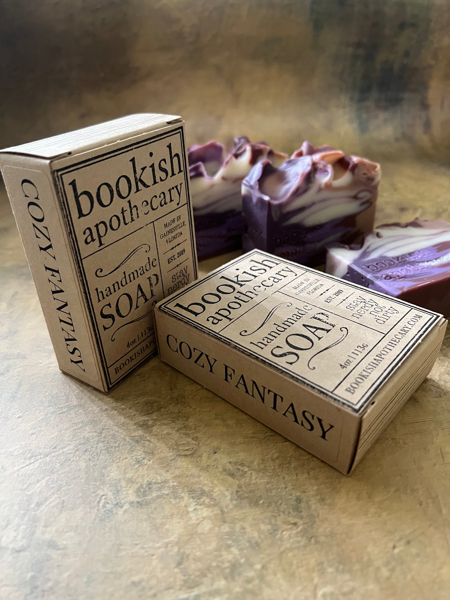Two rectangular boxes of Bookish Apothecary Cozy Fantasy Handmade Soap resting on a textured beige surface, with a purple and white patterned cloth in the background.