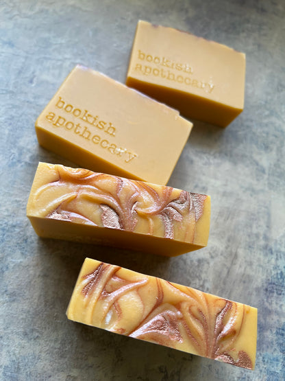 Four handmade rectangular bars of soap from Bookish Apothecary arranged diagonally on a textured gray slate surface. The soap bars feature a warm golden-yellow base with one bar displaying a metallic copper swirl pattern, and the others showing the embossed brand name.