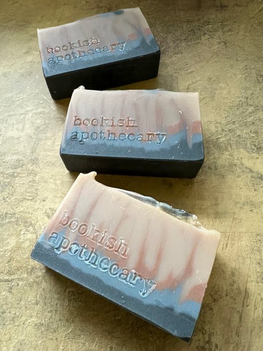 Three handmade soap bars with a marbled gray, light gray, and tan design stamped with the brand name 'bookish apothecary', displayed on a textured beige surface.