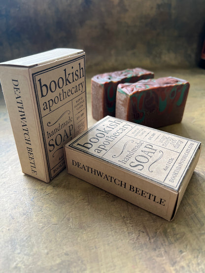 Two rectangular soap boxes labeled 'Bookish Apothecary Soap' and 'Deathwatch Beetle' are shown on a textured stone-like surface, with a stack of patterned soap bars in the background.