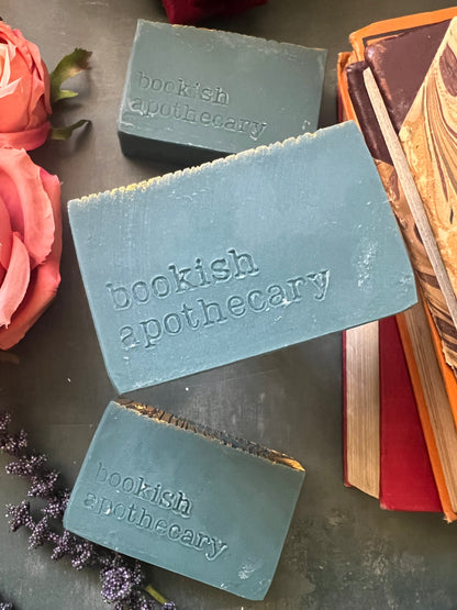 Three bars of soap with 'bookish apothecary' branding on a textured surface with flowers.