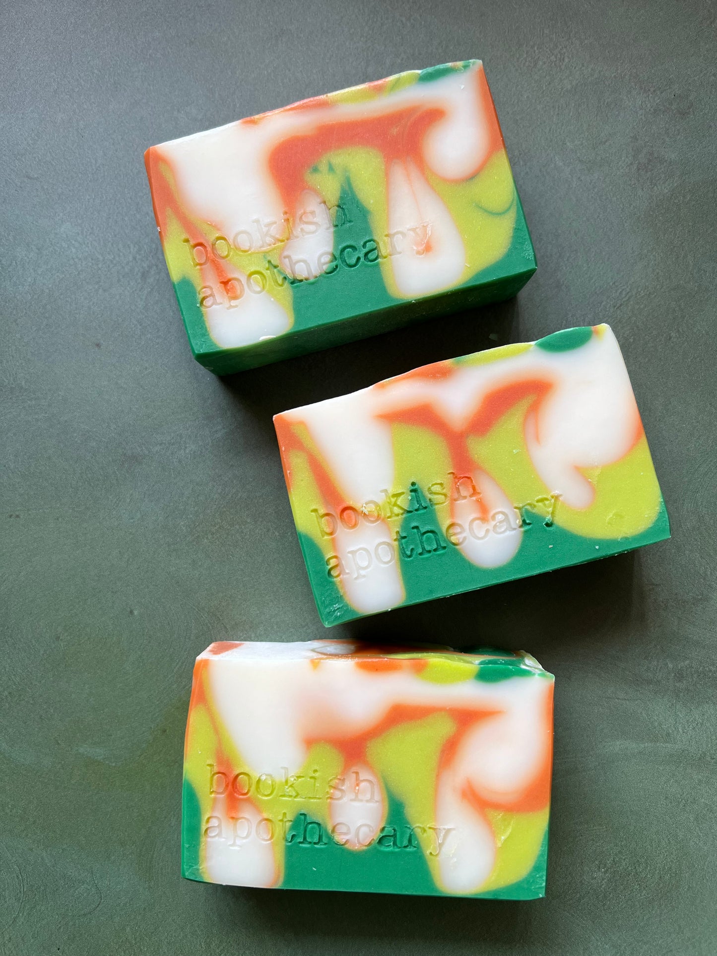 Three bars of swirled soaps comprised of orange, chartreuse and green soap stamped with bookish apothecary branding.