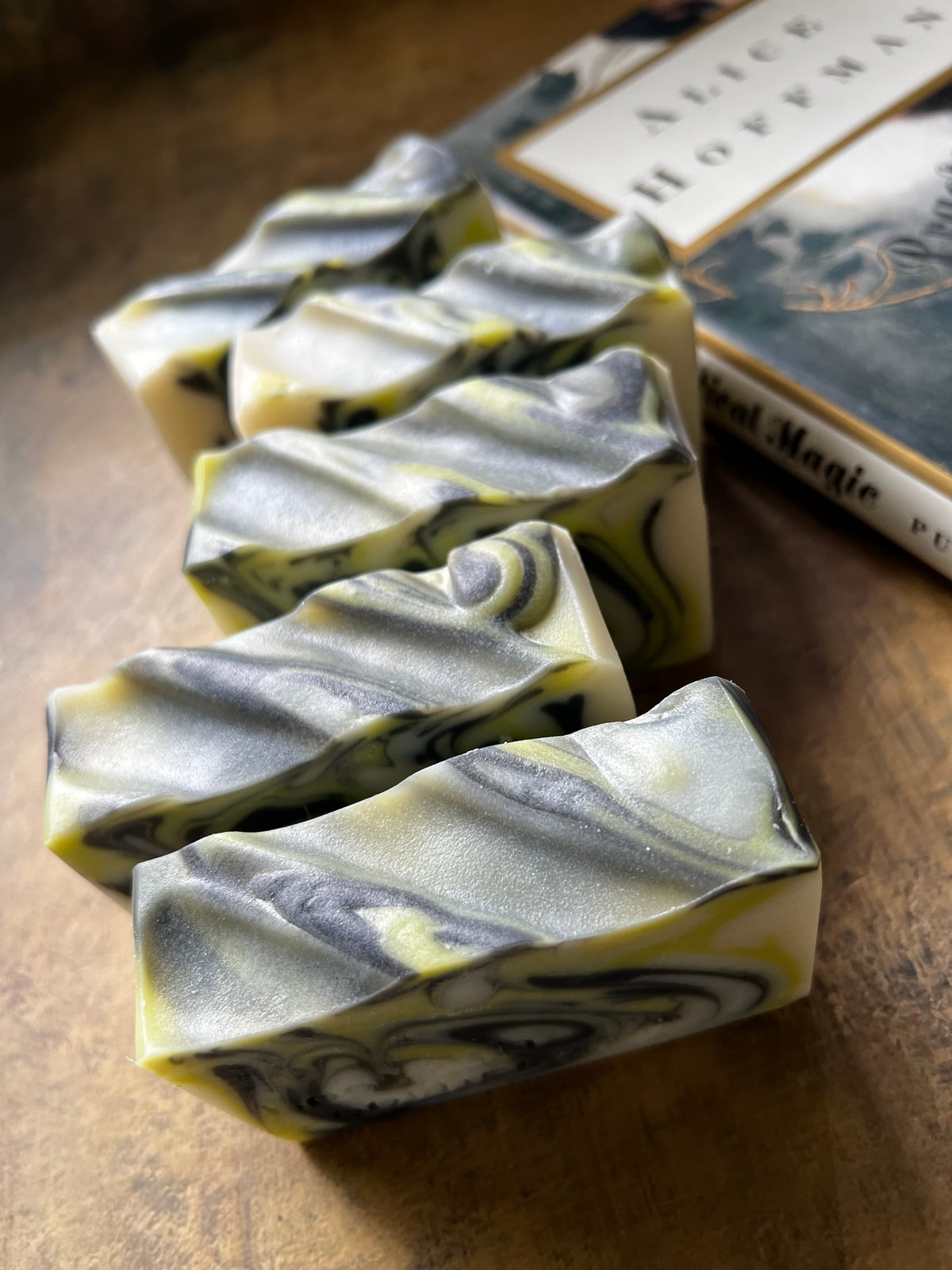Five marbled soap bars with a white, gray, and yellow swirl pattern arranged on a rustic wooden surface next to a book.