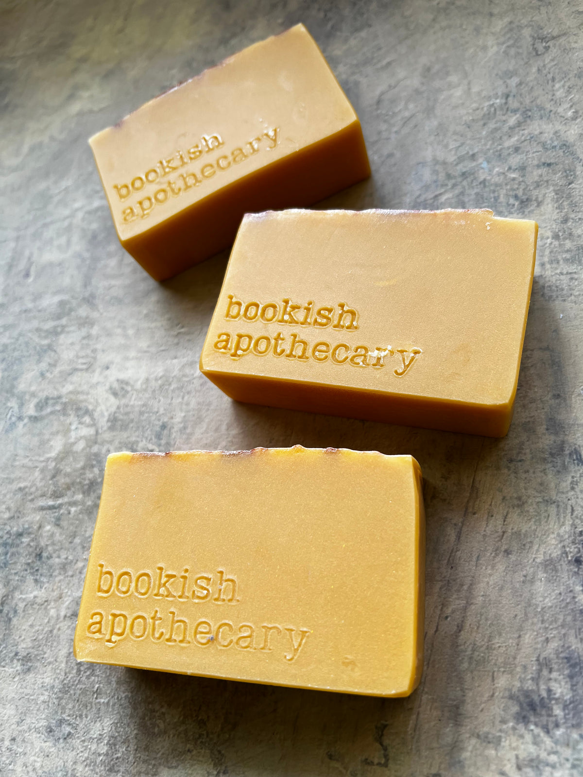 Three rectangular bars of handmade soap in a warm golden-yellow color, embossed with the brand name 'bookish apothecary', arranged diagonally on a textured gray slate surface.