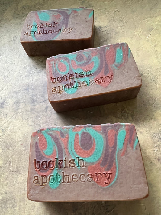 Three rectangular handmade soap bars with a marbled pattern of teal, pink, and brown, each stamped with the brand name 'bookish apothecary', arranged on a textured beige surface.
