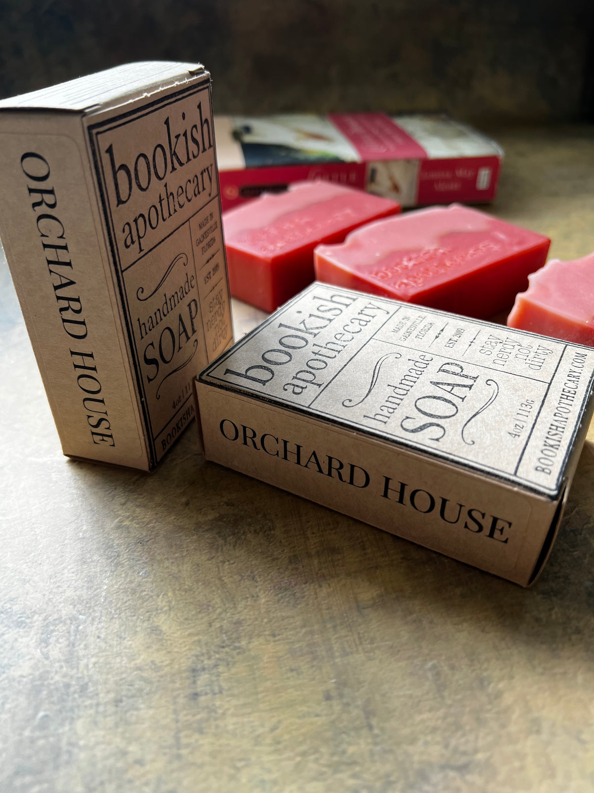 Several pink rectangular bars of soap and matching paperboard boxes printed with the words 'bookish apothecary' and 'ORCHARD HOUSE' are arranged on a textured tabletop.