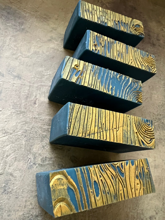 Five dark green rectangular of handmade soap  with a gold wood-grain textured top, arranged diagonally on a textured gray concrete surface.