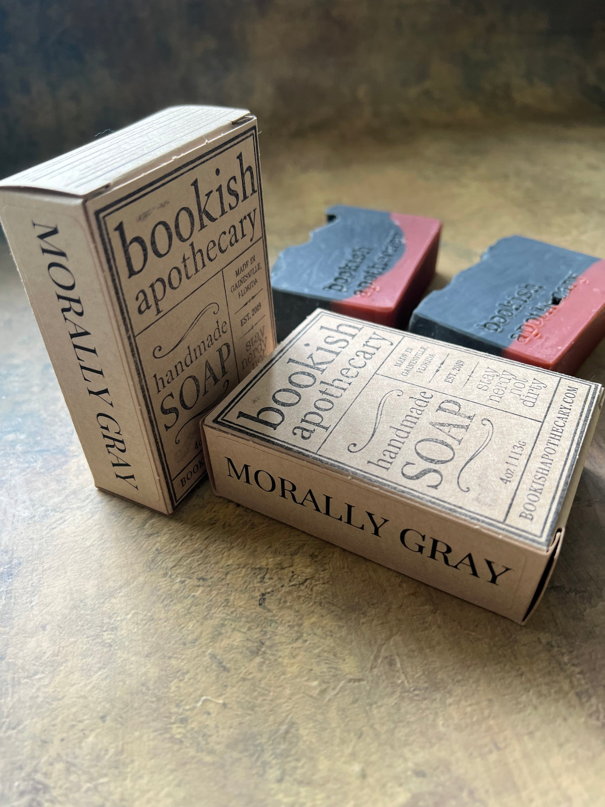 Two vintage-style soap bars in matching rectangular boxes labeled 'Bookish Apothecary' and 'Morally Gray,' arranged on a textured stone-like surface.