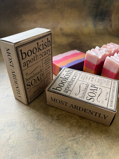 Two boxed bars of handmade soap from Bookish Apothecary on a textured stone surface, with additional pink, orange, and purple soap bars in the background.