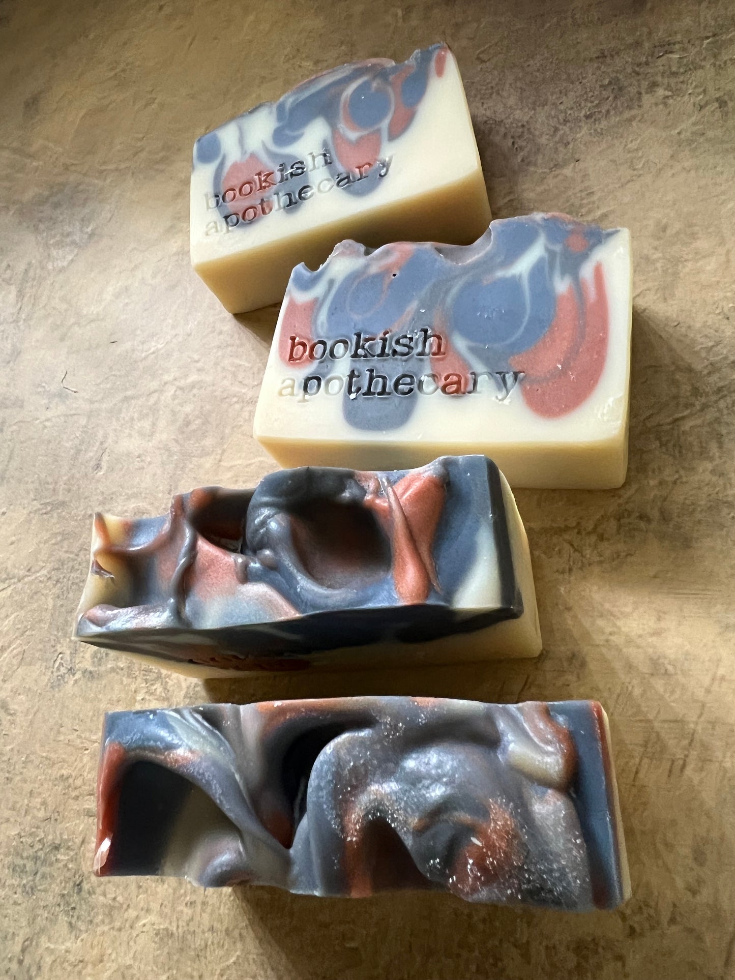 Four handmade artisanal soap bars with marbled swirls of cream, slate blue, and copper-brown, displayed on a textured beige surface.