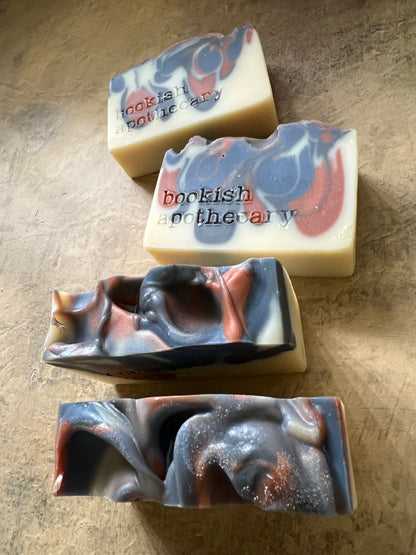 Four handmade artisanal soap bars with marbled swirls of cream, slate blue, and copper-brown, displayed on a textured beige surface.