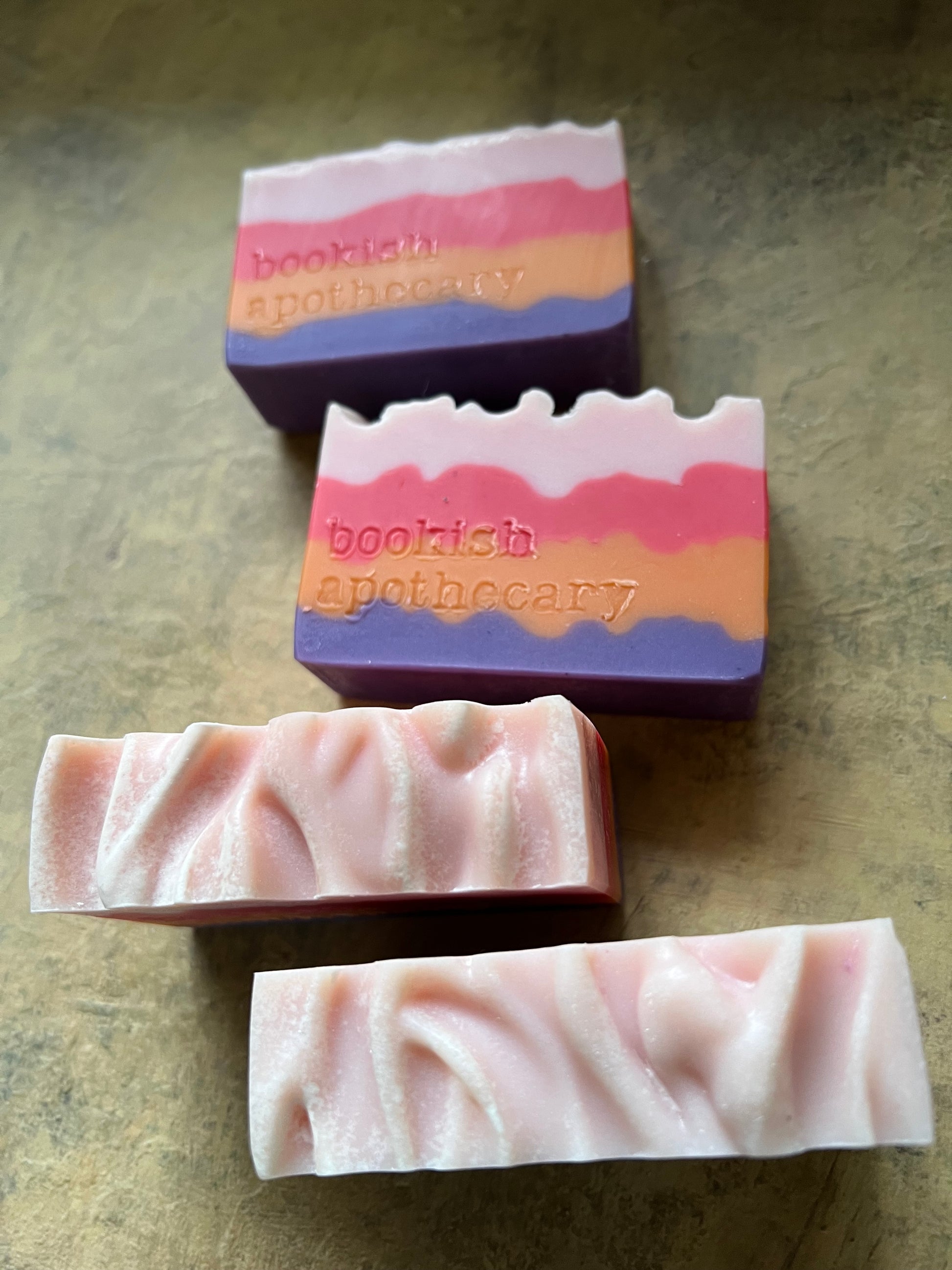 Four handmade layered soap bars with pink, orange, and purple stripes, stamped with the brand name 'bookish apothecary', displayed on a textured brown surface.