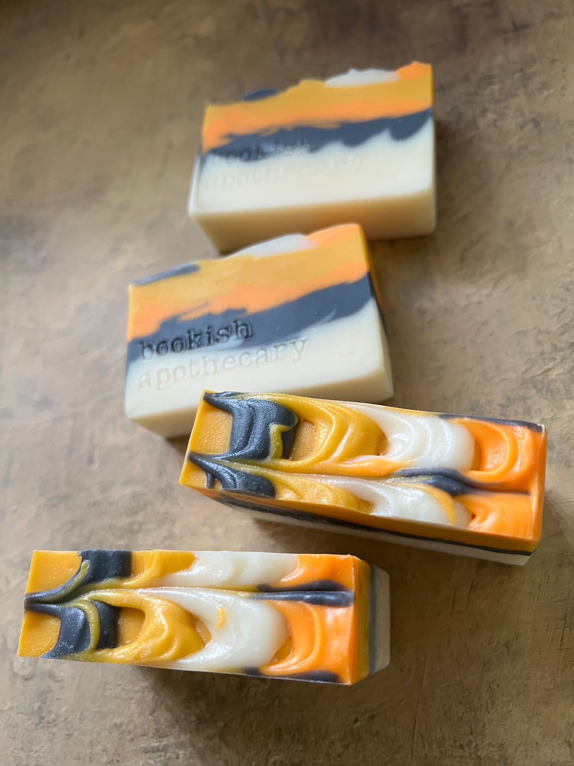 Four rectangular handmade soap bars with a marbled swirl pattern of orange, cream, white, and dark blue-gray, arranged on a textured brown surface.