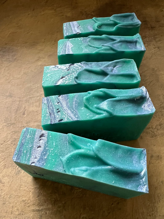 Five rectangular handmade soap bars with a green and blue marbled pattern and raised abstract wave-like details, arranged against a textured tan background.