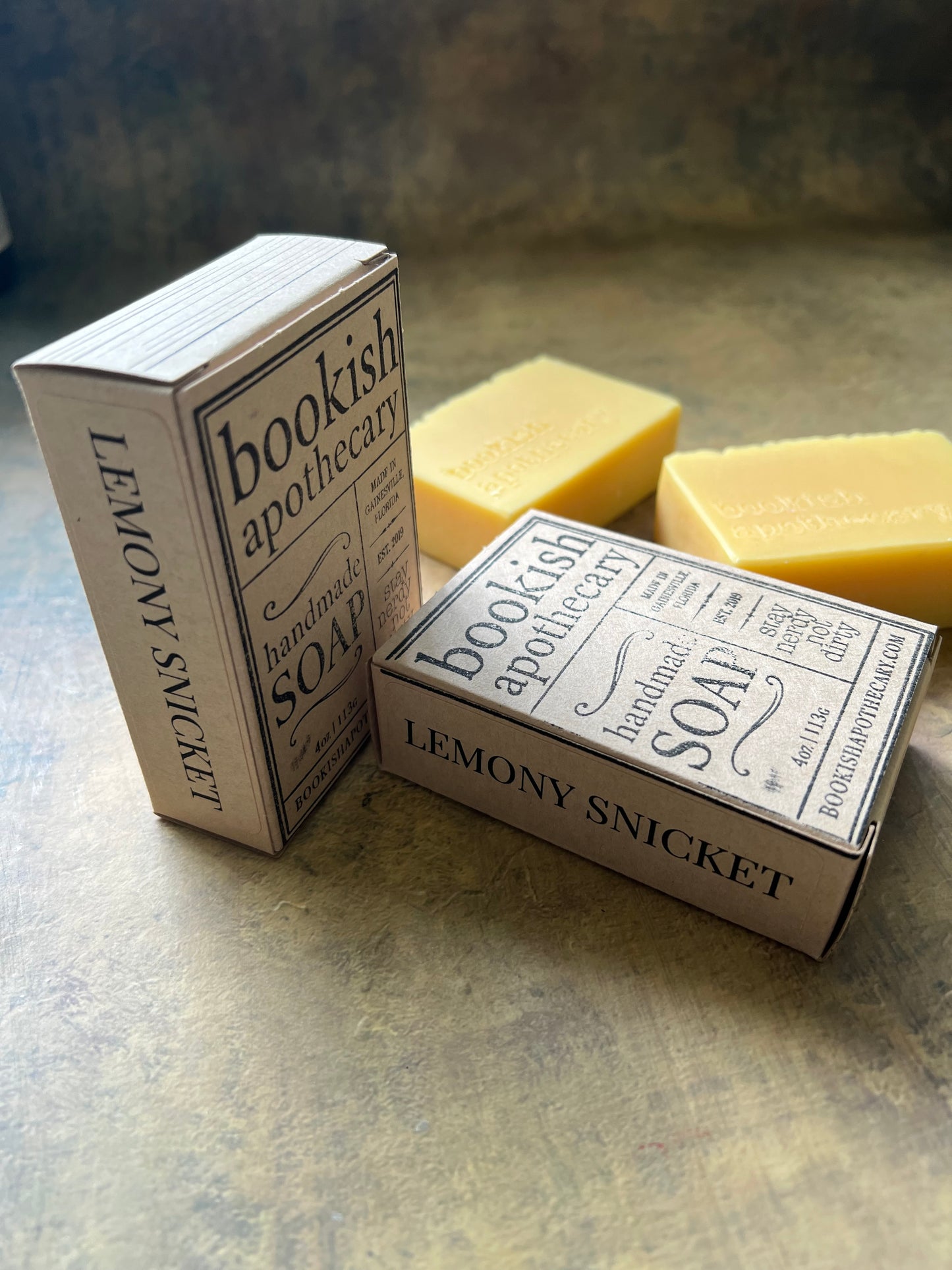 Two boxed bars of soap labeled 'Bookish Apothecary Lemony Snicket' with pale yellow soap bars visible behind them, photographed on a textured gray surface.
