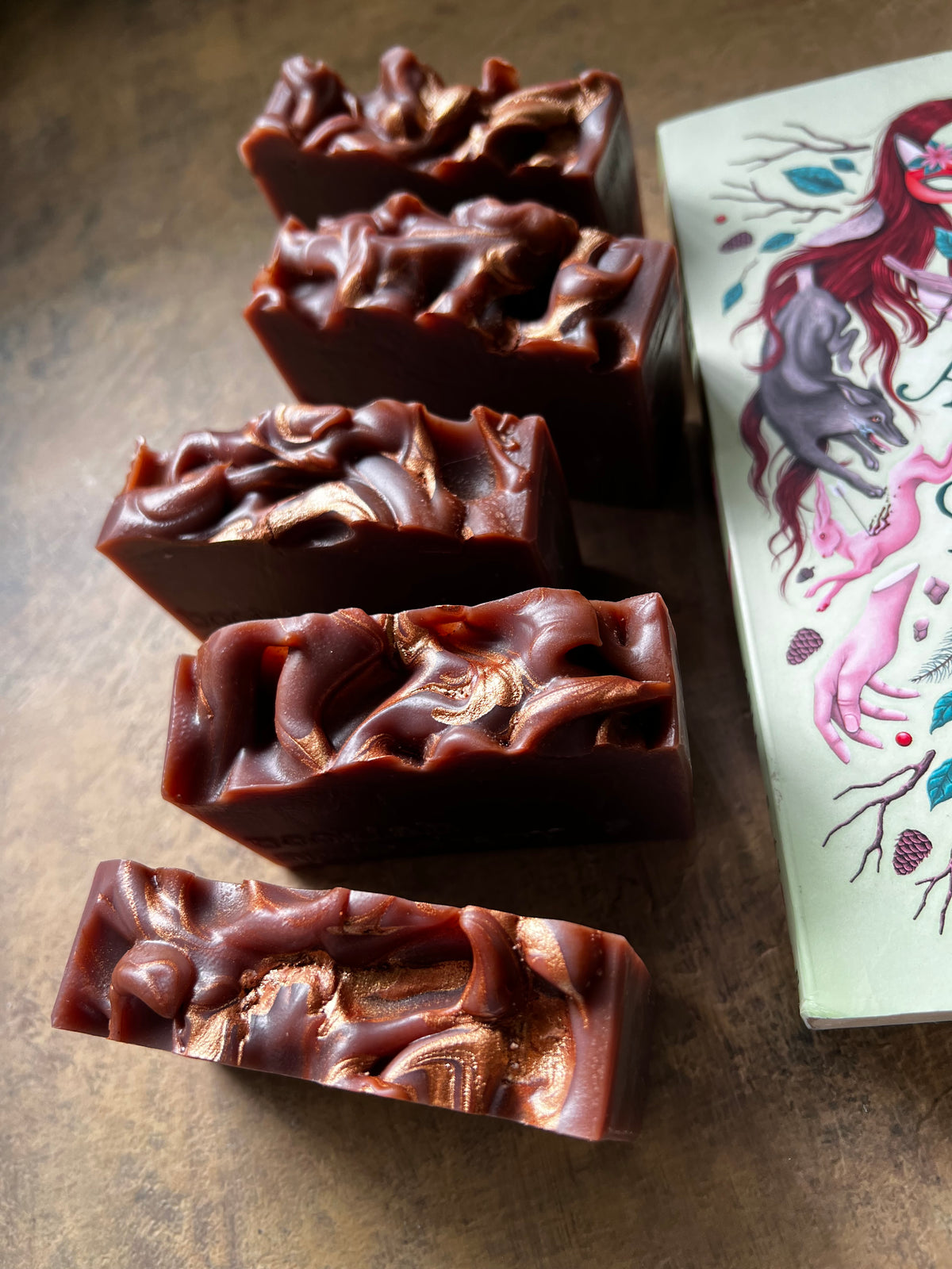 Five rectangular bars of handmade soap with a glossy, textured dark reddish-brown finish, arranged diagonally on a dark brown surface next to a decorative illustrated book.