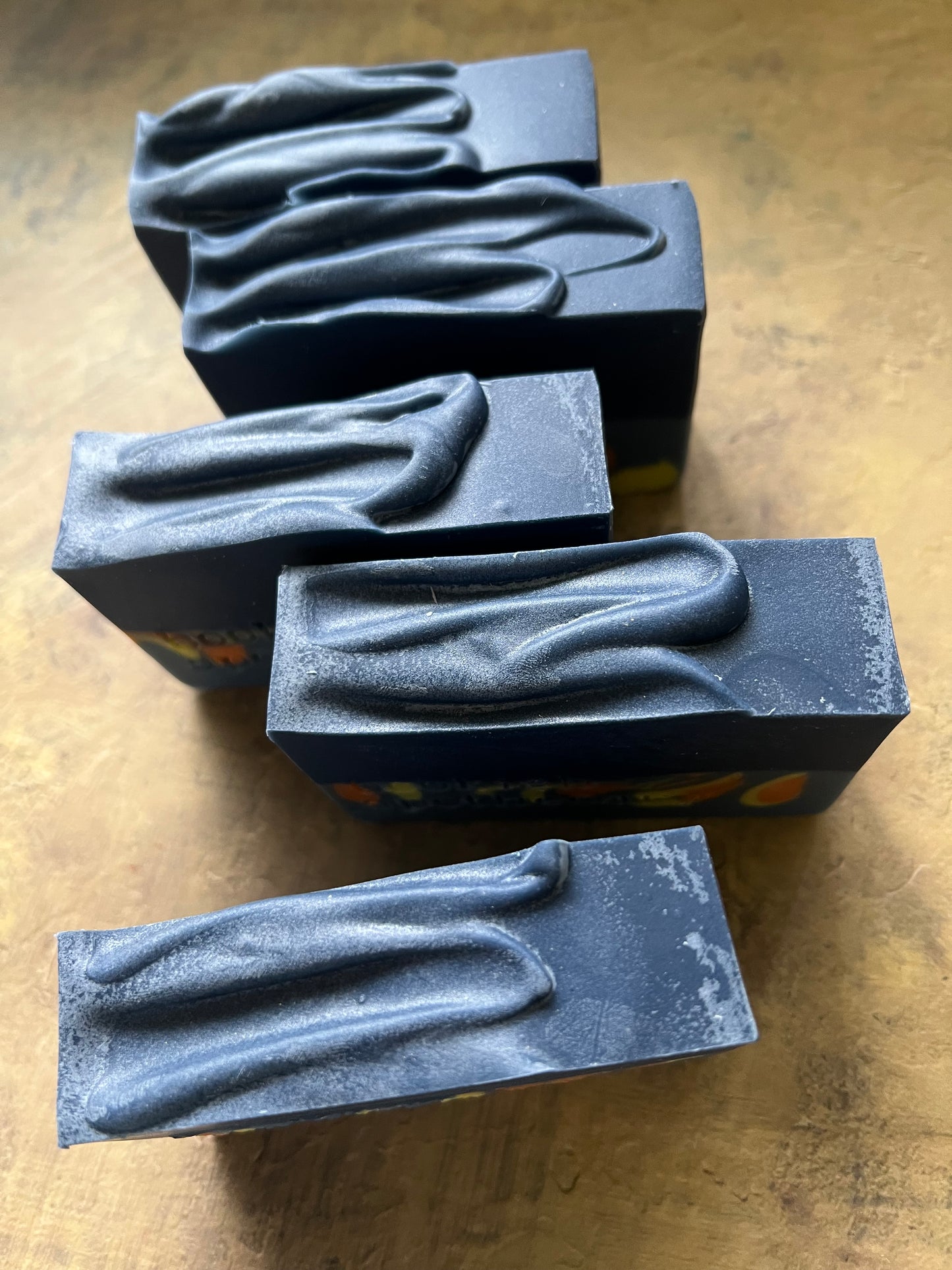 Five rectangular dark blue soap bars with abstract wave-like patterns, arranged on a textured tan surface.