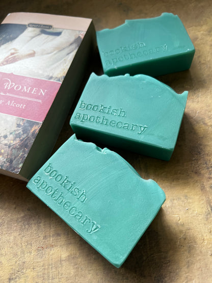 Three green soap bars embossed with 'bookish apothecary' are arranged on a wooden surface next to a book titled 'Little Women'.