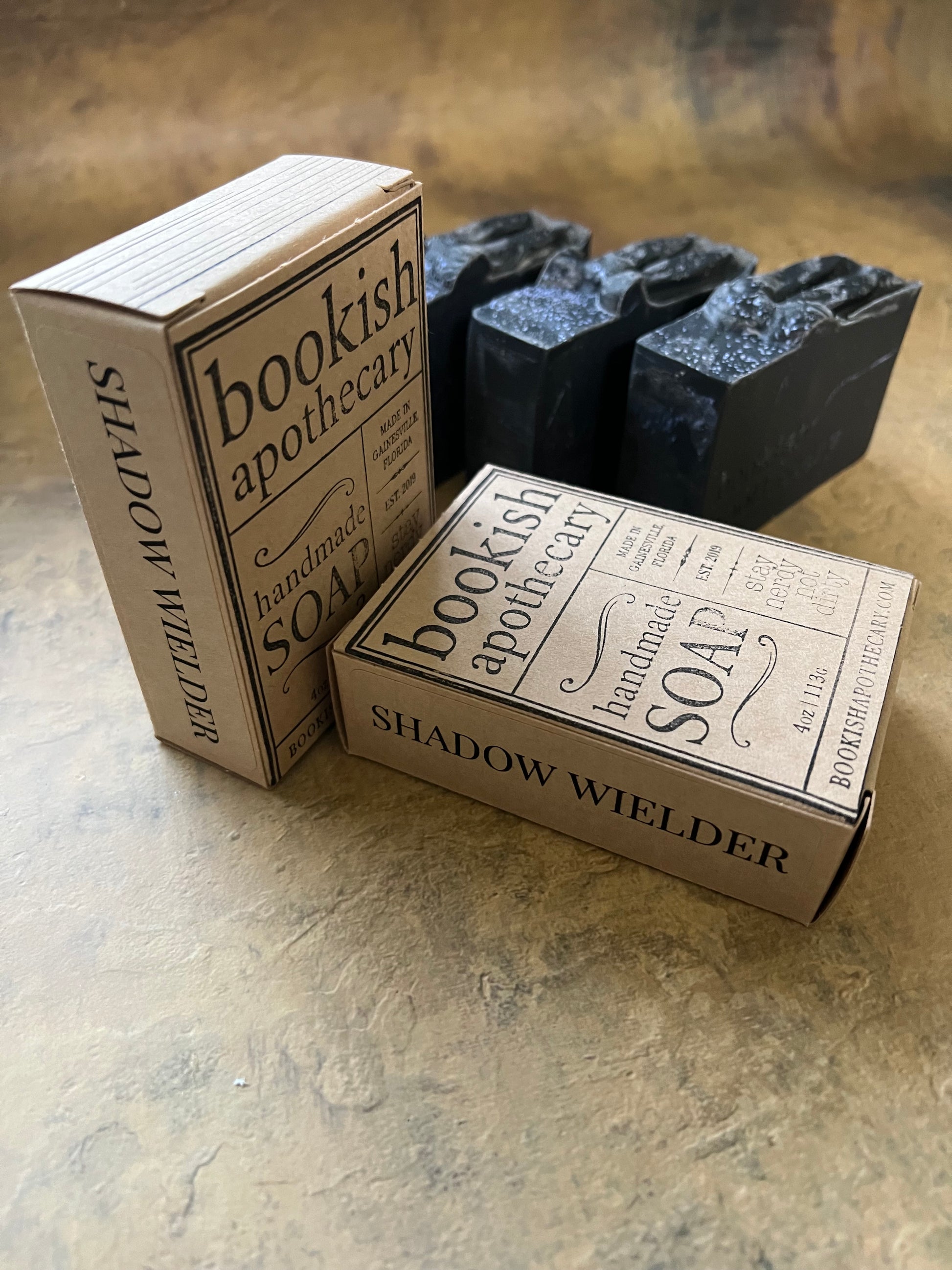 Two vintage-style cardboard apothecary soap boxes stacked on top of each other, with three dark rectangular bars of soap resting behind them, photographed on a textured beige surface.