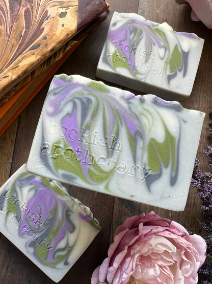 Three marbled soap bars with a brand name on a wooden surface, accompanied by a pink flower.