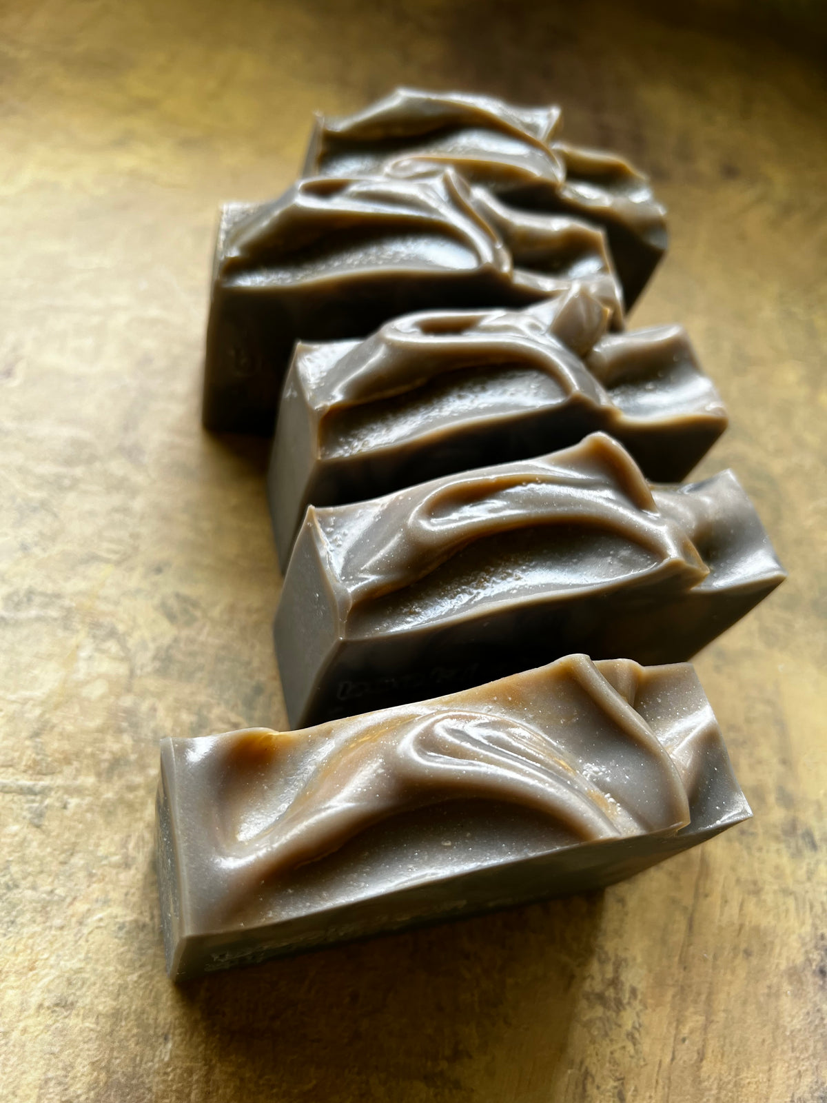 Five rectangular bars of handmade soap with textured, swirled tops, arranged diagonally on a rustic, warm brown surface.