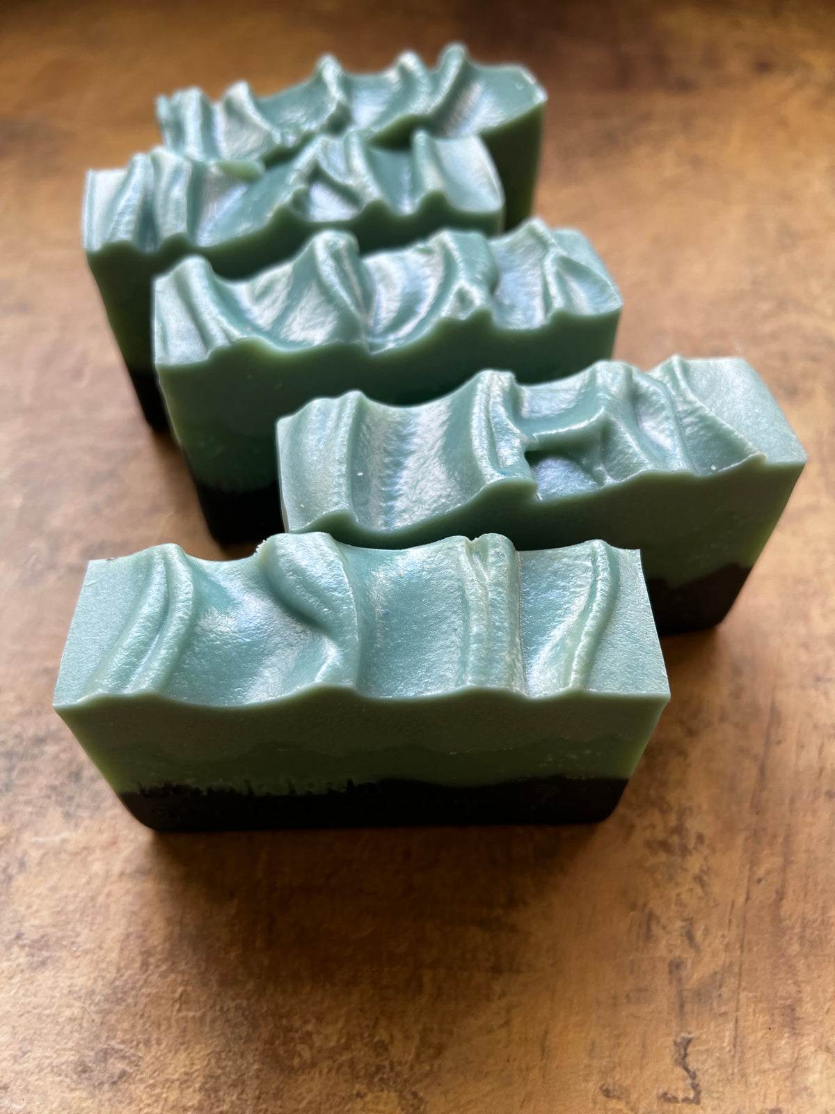 Four rectangular bars of handmade soap with a wavy, layered top and a dark base, arranged diagonally on a rustic wooden surface.