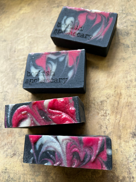 Four rectangular bars of handmade soap with a marbled pattern of black, red, pink, and white, arranged diagonally on a rustic wooden surface. The top two bars are embossed with the brand name 'borisfish apothecary'.