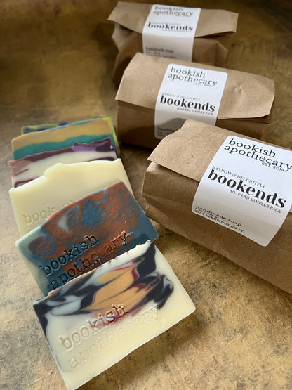Several handmade soap bar ends with marbled cream, black, blue, and orange swirls are arranged on a textured brown surface, with multiple brown kraft paper bags wrapped in white labels reading 'bookish apothecary bookends' behind them.