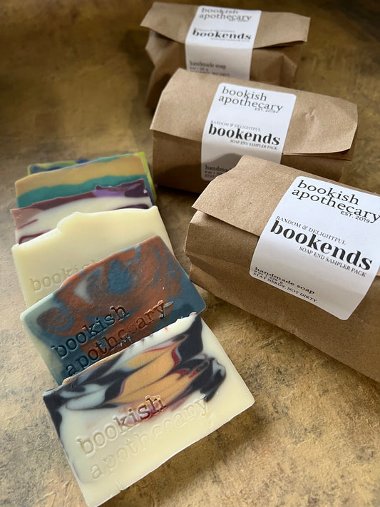 Several handmade soap bar ends with marbled cream, black, blue, and orange swirls are arranged on a textured brown surface, with multiple brown kraft paper bags wrapped in white labels reading 'bookish apothecary bookends' behind them.
