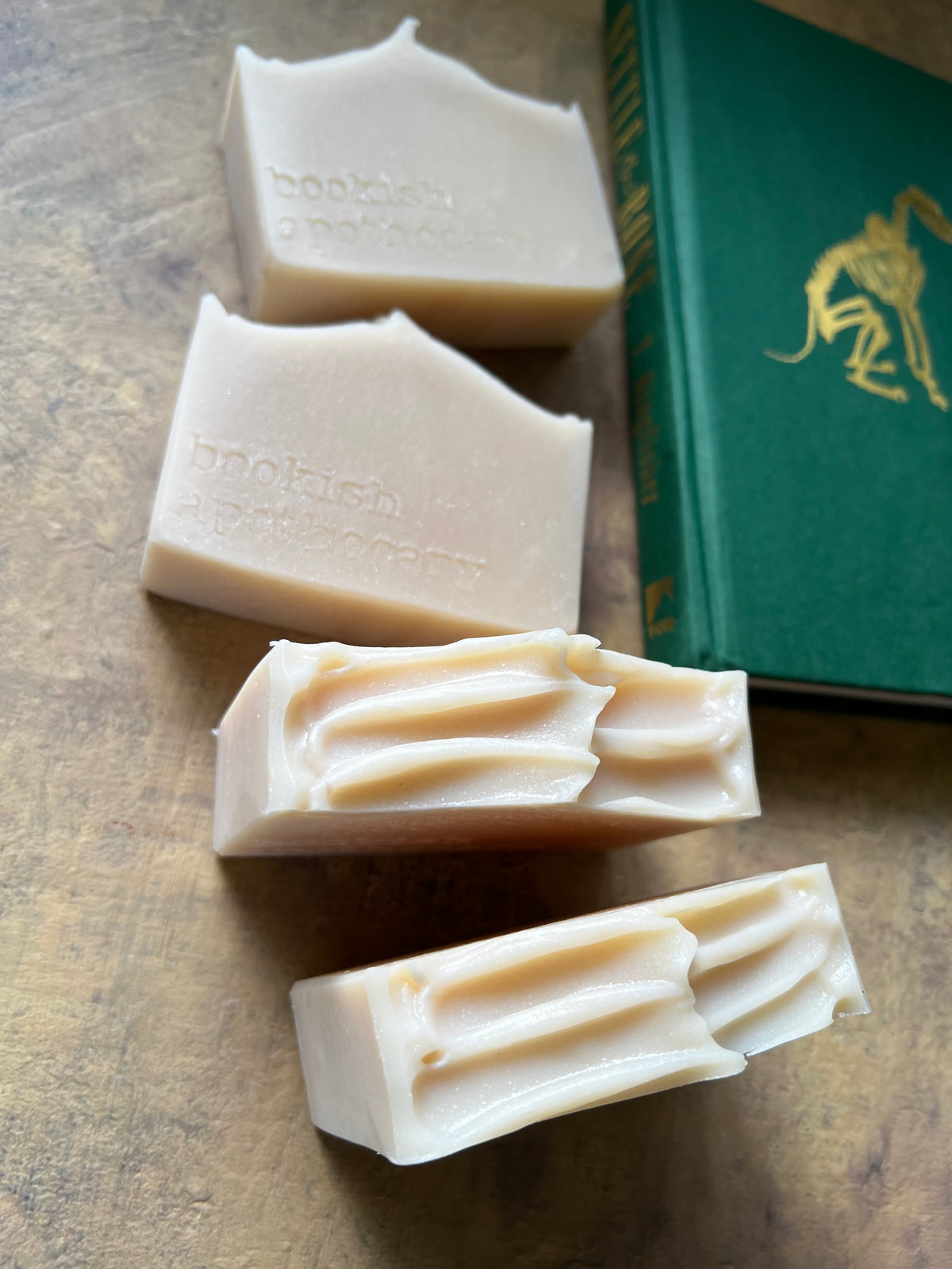 Four bars of handmade soap in a warm cream-beige color, arranged diagonally on a textured brown surface next to a green book with gold lettering.