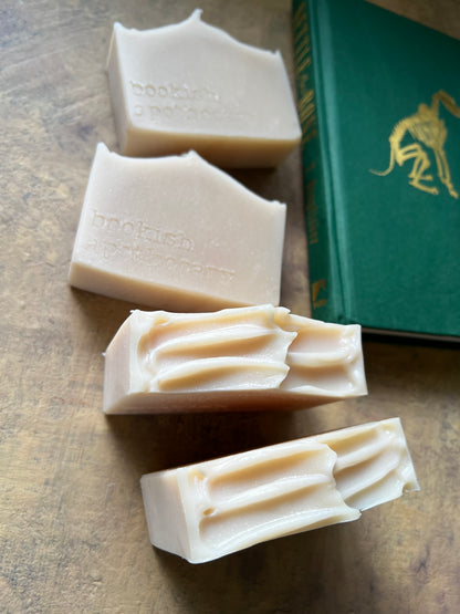 Four bars of handmade soap in a warm cream-beige color, arranged diagonally on a textured brown surface next to a green book with gold lettering.