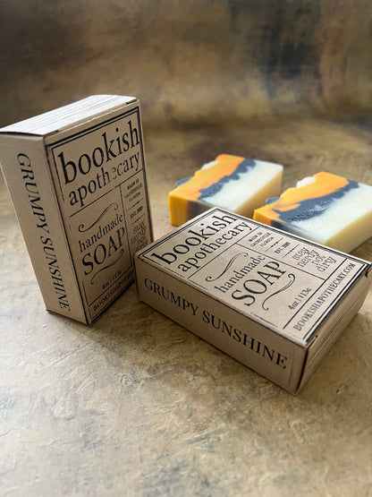 Two boxed bars of soap labeled 'Grumpy Sunshine' and 'Bookish Apothecary Soap' displayed on a textured stone surface, with one bar partially visible behind the boxes.