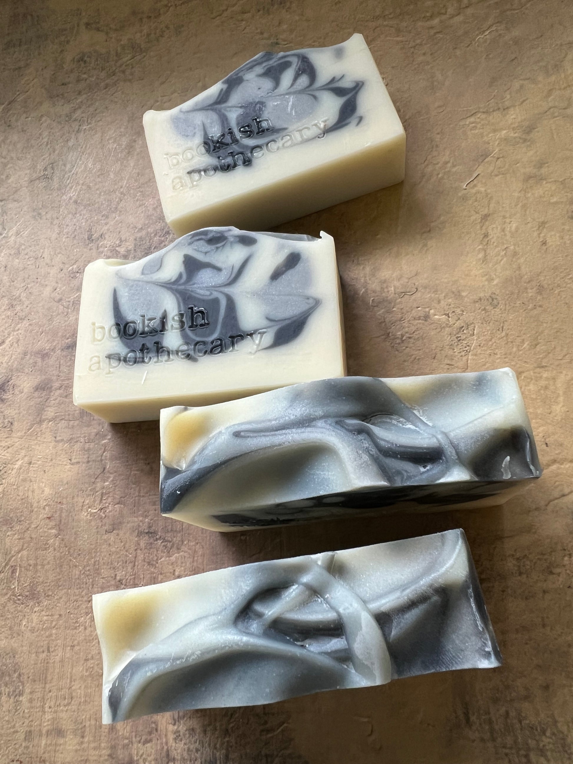 Four handmade soap bars with a marbled cream and dark gray swirl pattern, arranged on a textured brown surface. The brand name 'bookish apothecar' is stamped on  the soaps.