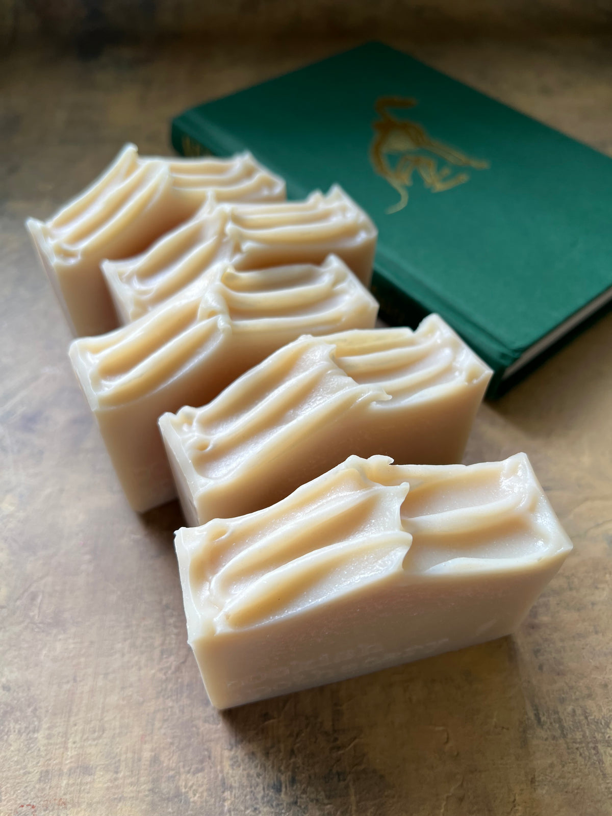 Four rectangular bars of creamy off-white soap with ridged tops, arranged diagonally on a wooden surface next to a green book.