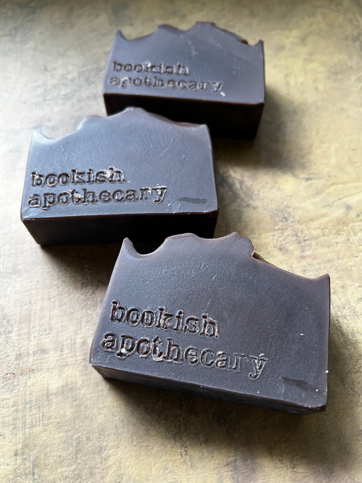 Three dark brown rectangular bars of soap with a wavy top edge, each embossed with the brand name 'bookish apothecary', arranged on a textured metallic gold surface.