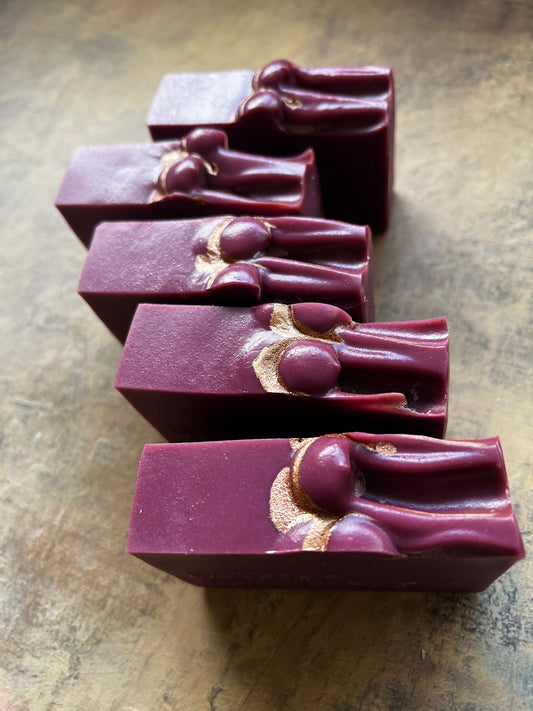 Five rectangular bars of handmade soap in a deep burgundy color, arranged diagonally on a textured brown surface. Each bar features a raised decorative design of a stylized figure in the center with a gold metallic inlay.