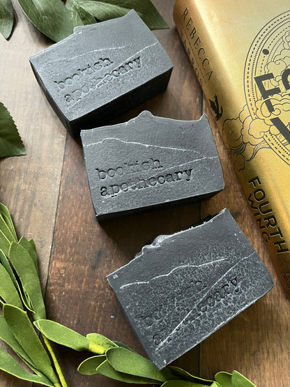 A bar of handmade soap with a gray color and speckled with dark dots, placed on a wooden surface with green leaves in the background and a label that reads 'COLLECTOR EDITION online exclusive'.