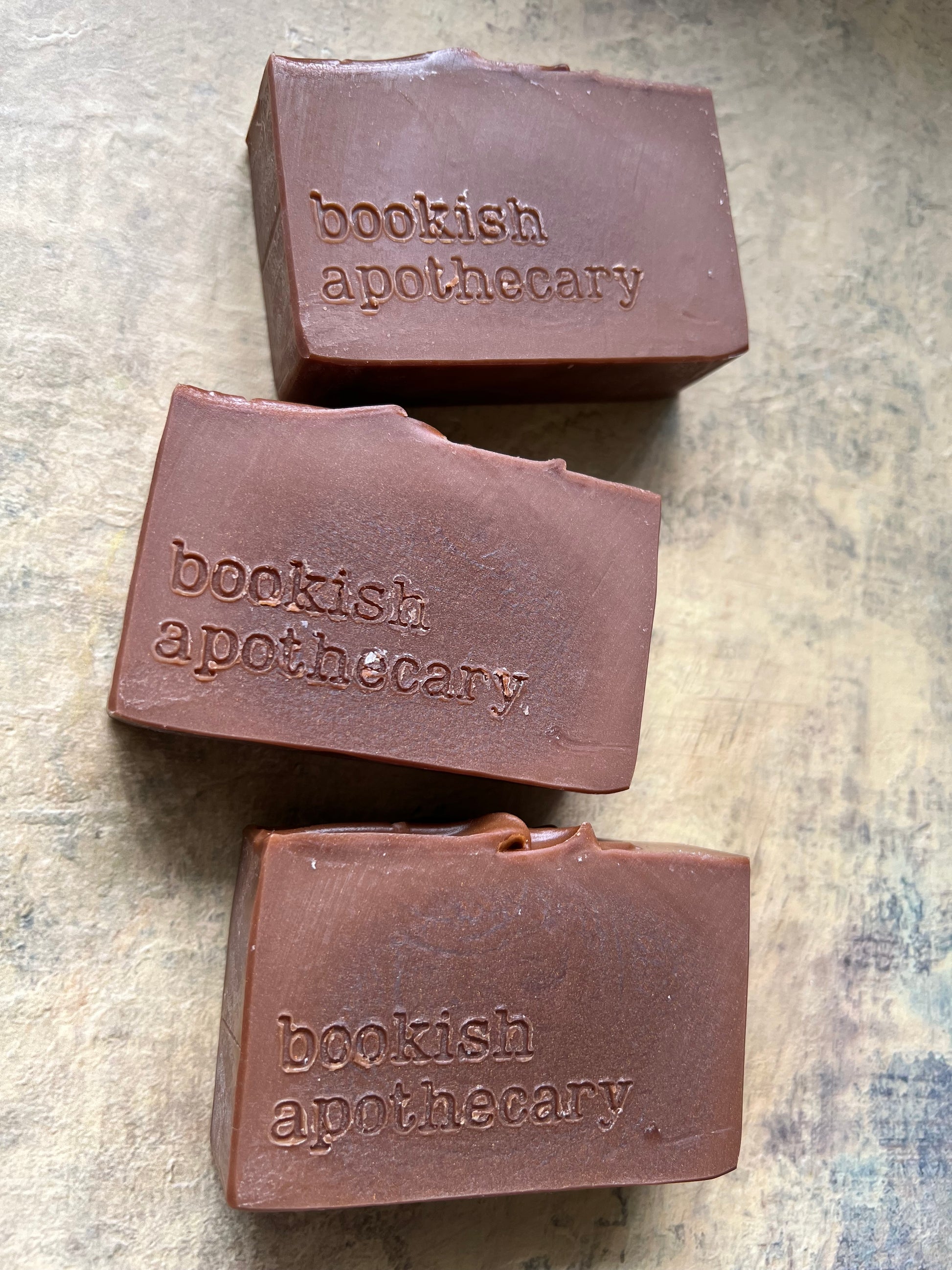 Three rectangular bars of brown soap with the brand name 'bookish apothecary' embossed on the front, arranged on a textured beige surface.