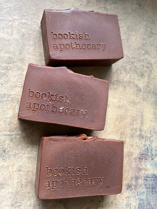 Three rectangular bars of brown soap with the brand name 'bookish apothecary' embossed on the front, arranged on a textured beige surface.