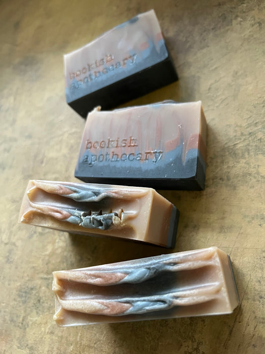 Four handmade artisanal soap bars with marbled beige, gray, and black patterns, arranged on a textured beige surface. Two bars feature the brand name 'bookish apothecary' stamped on the front
