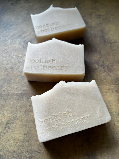 Three handmade soap bars with a rustic, hand-crafted look, embossed with the brand name 'bookish apothecary', arranged diagonally on a textured brown surface.