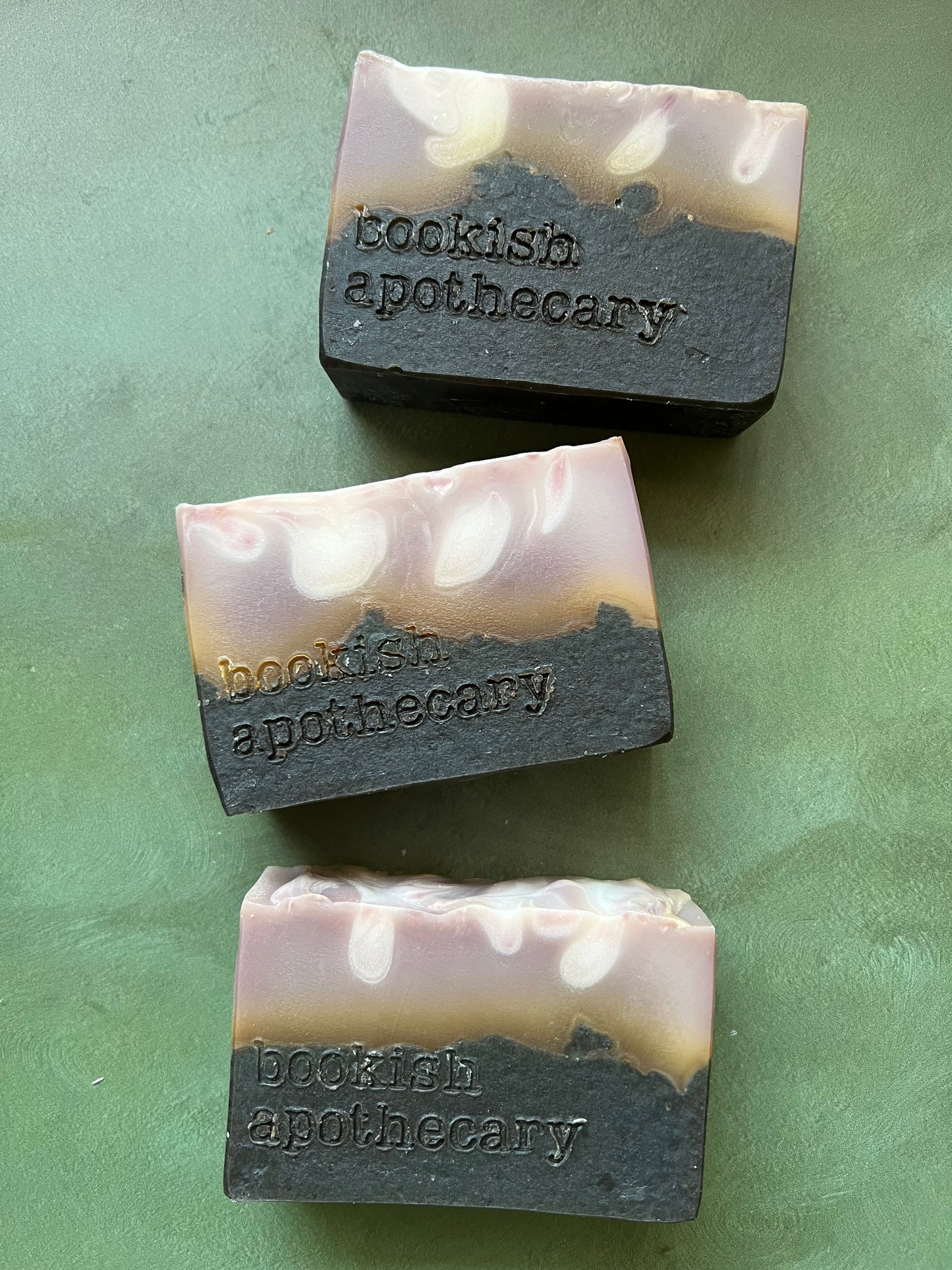 Three bars of  soap with a tan top and black bottom stamped with bookish apothecary branding.