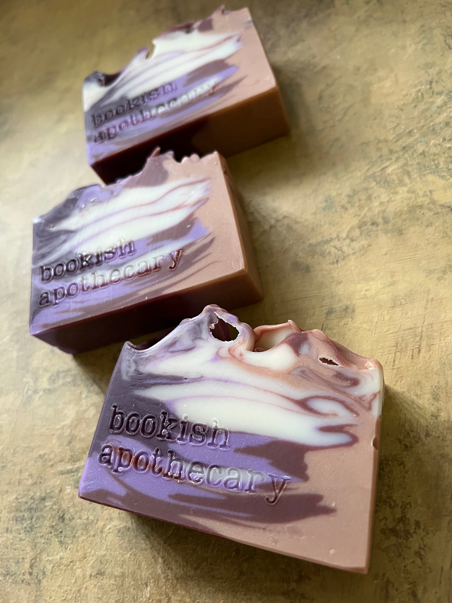 Three handmade artisanal soap bars with a swirled lavender, purple, and white pattern, stamped with the brand name 'bookish apothecary', arranged diagonally on a textured gold surface.