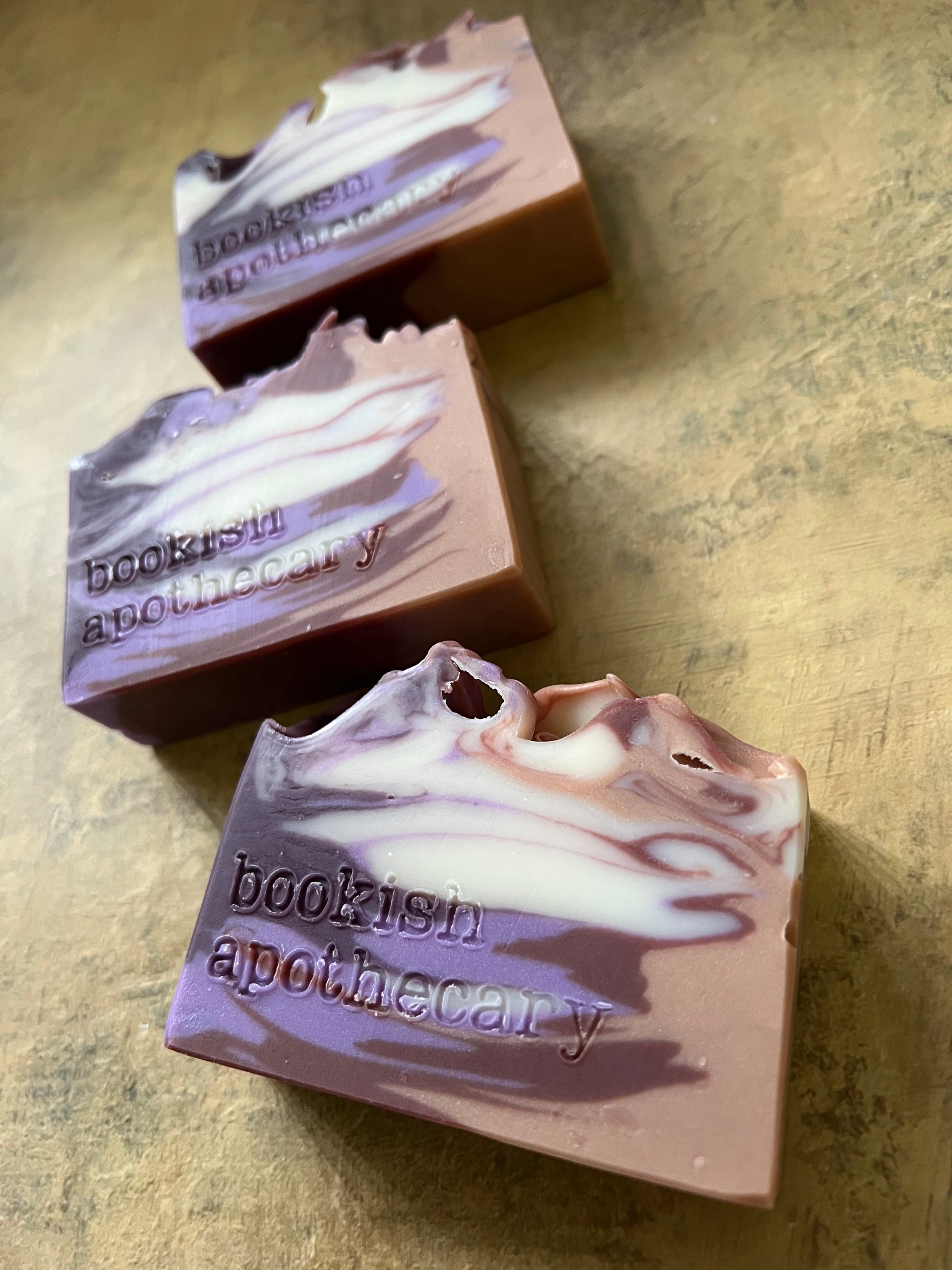 Three handmade artisanal soap bars with a swirled lavender, purple, and white pattern, stamped with the brand name 'bookish apothecary', arranged diagonally on a textured gold surface.