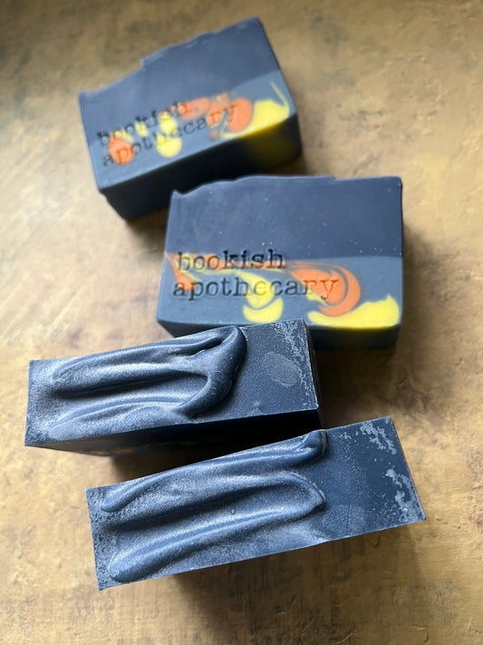 Four rectangular bars of soap with dark blue-gray bodies and yellow-orange swirls, featuring the brand name 'bookish apothecary' stamped on them, arranged on a textured brown surface.